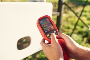 Technician using a handheld thermal imaging camera to detect abnormal heat patterns in outdoor equipment.